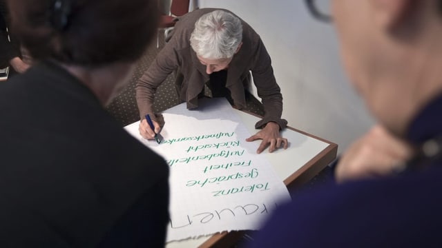 Eine ältere Frau schreibt etwas auf ein Papier eines Flipcharts.