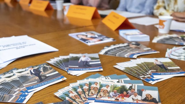 Werbeflyer liegen auf einem Tisch, im Hintergrund Menschen mit Namensschildern