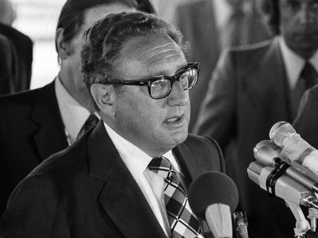 Ein Foto in schwarz/weiss von Henry Kissinger. Er spricht in verschiedene Mikrofone.