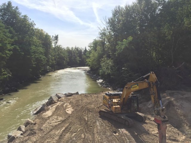 Bagger auf Aufschüttung in Fluss, rundherum Bäume.