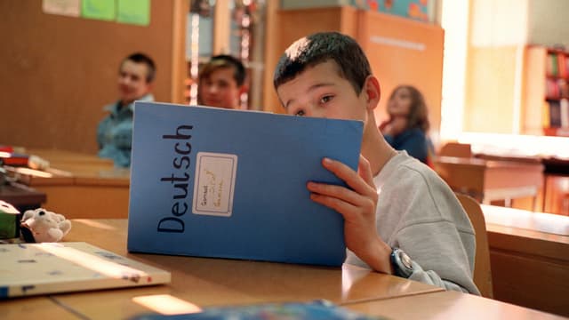 Ein Junge schaut in einer Schulklasse konzentriert in ein blaues Deutsch-Heft. 