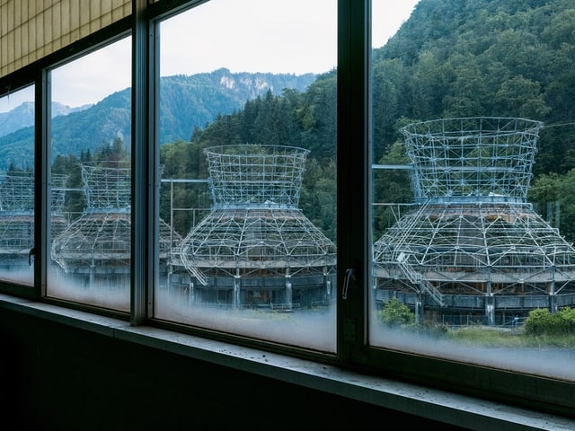 Blick aus dem Fenster auf Industriebauten vor bewaldeten Bergen.