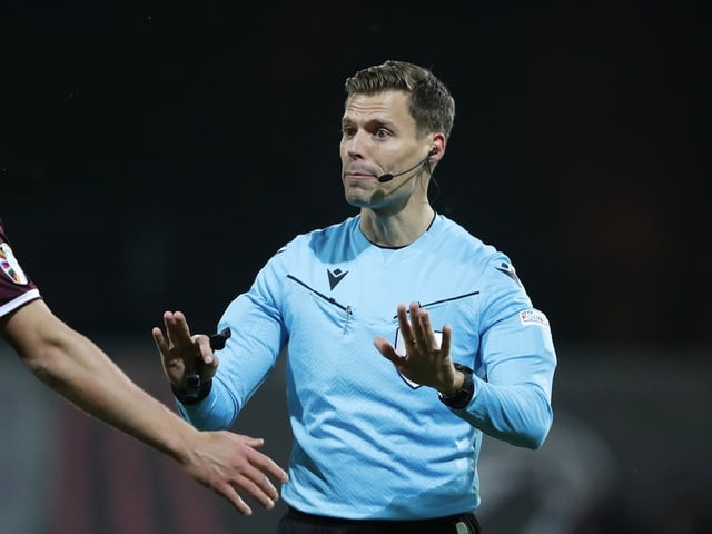 Schiedsrichter in hellblauer Uniform auf einem Fussballfeld mit erhobenen Händen.