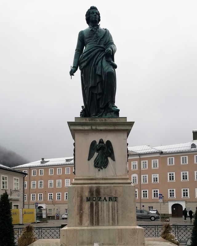 Eine Statue eines Mannes. Unten ein Engel. Im Hintergrund ein altes, grosses Gebäude.