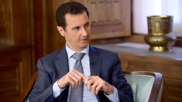 Assad sitzt in Anzug und Krawatte in einem Sessel.