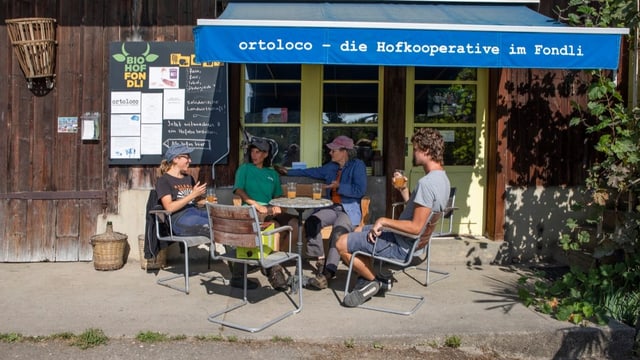 Vier Menschen sitzen vor einer alten Hofwand um einen Tisch, auf Sonnenschutz steht «ortoloco».