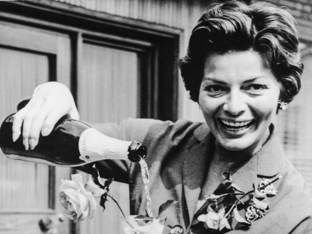 Lys Assia hebt eine Flasche Champagner und füllt ein Glas.