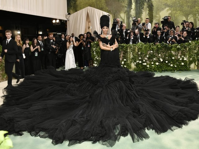 Frau in üppigem schwarzen Kleid auf rotem Teppich, von Fotografen umgeben.