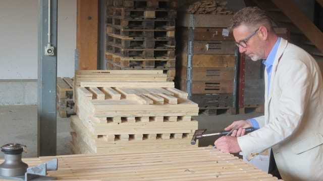 Mann in beigem Jacket hält Hammer über Holzpallett.