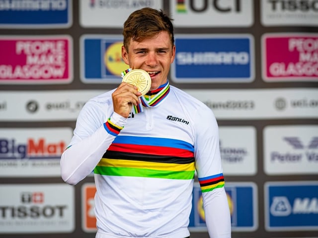 Radfahrer mit Regenbogentrikot beisst auf Medaille auf dem Podium.