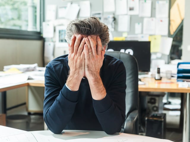 Gestresster Mann hält seinen Kopf in den Händen in einem Büro.