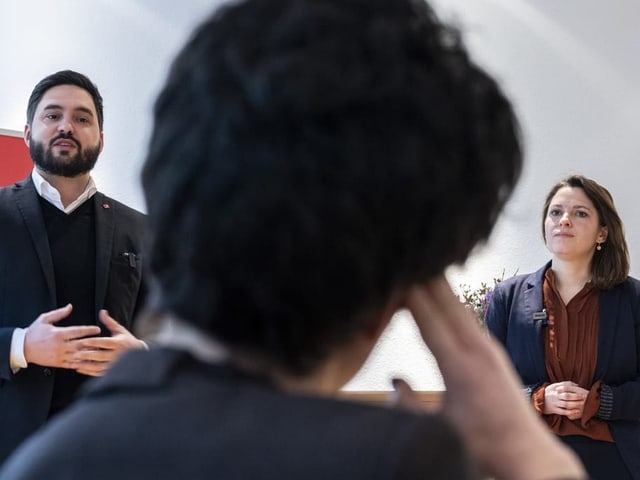 Mann mit Bart und Anzug und Frau mit schulterlangen Haaren und Anzug im Hintergrund, davor verschwommen eine Person