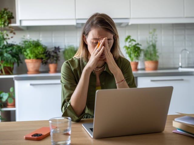 Eine Frau greift sich in einer Küche sitzend vor dem Computer ins Gesicht und scheint sich die Augen zu reiben.