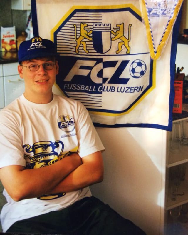 Ein junger Bursche mit Baseballmütze und weissem T-Shirt sitzt vor einem Tuch mit dem aufgedruckten Logo des FC Luzern.