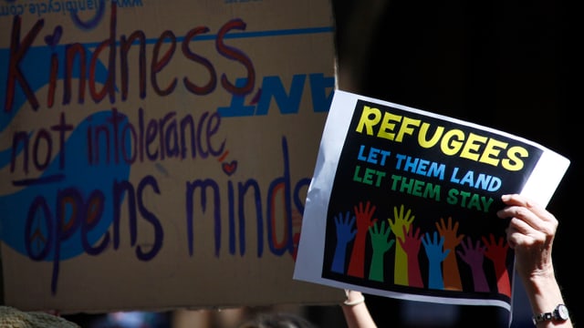 An einer Demo hält jemand ein Schild in die Höhe, darauf steht "Refugee, let them land, let them stay".