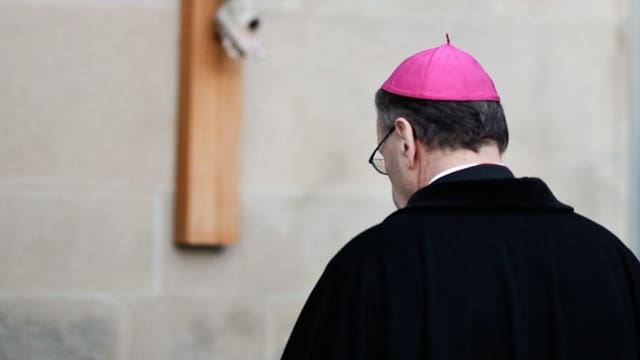 Der Churer Bischof Vitus Huonder von hinten mit einem purpurnen Pileolus vor einem Kreuz.