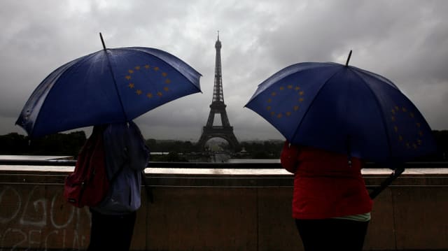 Zwei Personen unter blauen Regenschirmen schauen in Richtung des Eiffelturms.