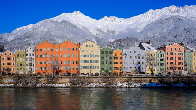 Häuser mit bunten Fassaden an einem Fluss. Im Hintergrund schneebedeckte Berge bei blauem Himmel