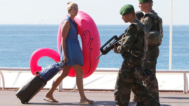 Zwei patrouillirende Soldaten an der Promenade von Nizza kreuzen eine sommerlich gekleidete Touristin mit Rollkoffer