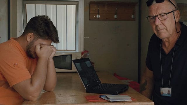 Links junger Mann in oranger Arbeiterkleidung, rechts älterer Mann mit Brille, beide am Tisch.