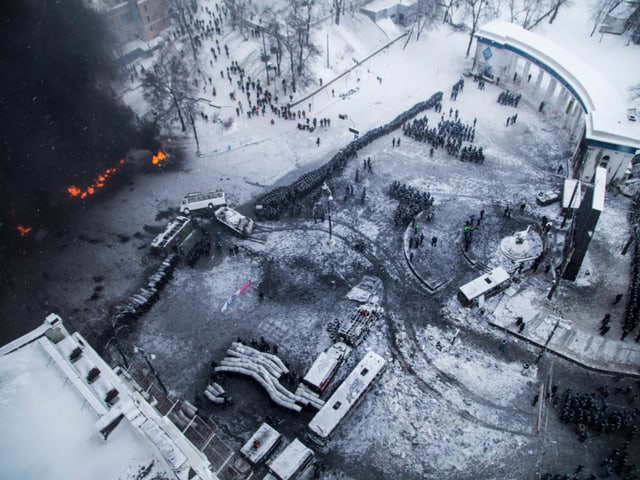 Aufnahme von oben: Auf einem verschneiten Kiewer Platz sieht man brenndende Barrikaden, ausgebrannte Autowracks und sich gegengüberstehende Polizisten und Demonstranten.