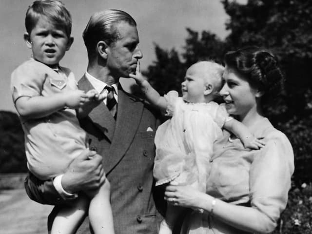 A boy is sitting in his father's arms, next to him is his mother in a dress holding a child in her arms.