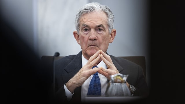 Jerome Powell hinter Schatten, die Hände in Merkel-Position vor sich.