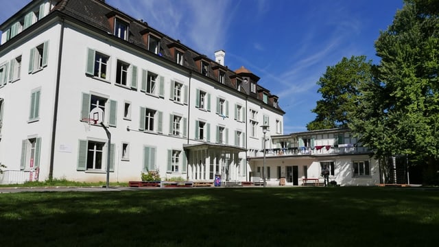 Ein grosses altes Haus. Davor ein grosser Rasenplatz und ein Basketballkorb.