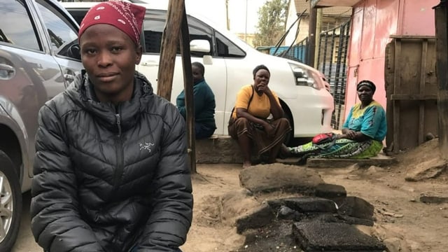 Hellen Achayo sitzt auf einem Autoreifen, im Hintergrund schauen drei Personen in die Kamera.