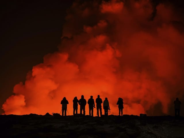 Die Silhouetten von sieben Personen sind vor rötlich aufsteigendem Rauch erkennbar.