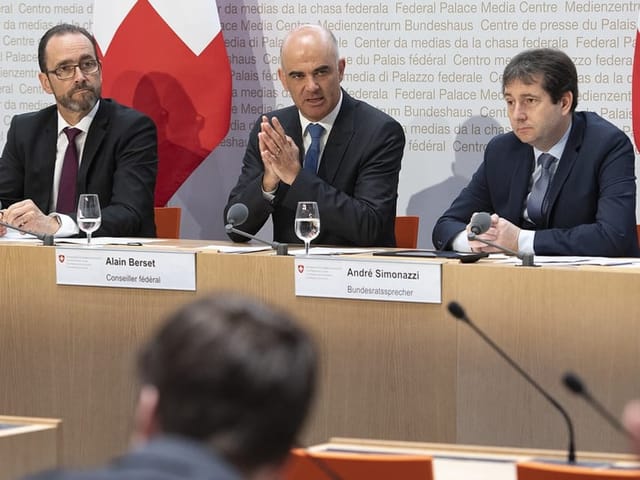 Drei Männer in Anzügen sitzen an einem Tisch an einer Medienkonferenz.