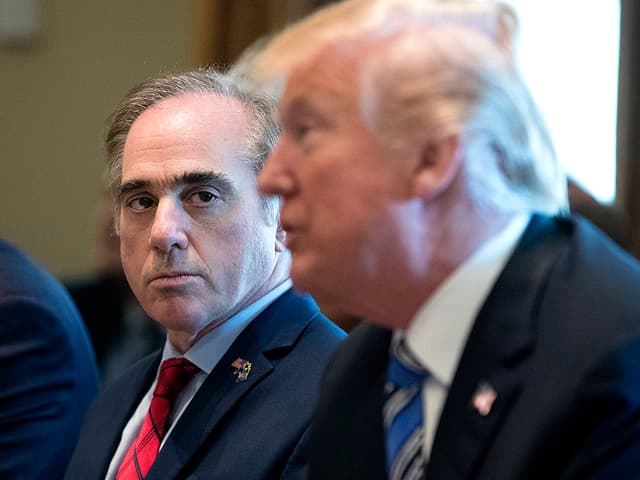 US-Präsident Donald Trump unscharf im Vordergrund. Hinter ihm, geschärft, der ehemalige Veteranenminister David Shulkin mit kühlem Blick.