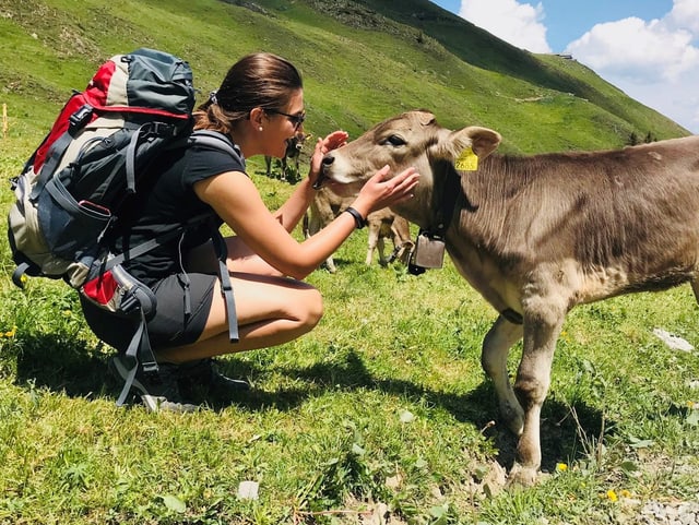 Angela auf einer Wiese mit Wanderrucksack. Sie hat gebeugte Knies und streichelt ein Kalb.