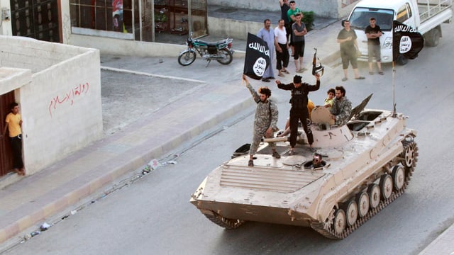 Junge Dschihadisten stehen auf einem rollenden Panzer und schwingen eine IS-Fahne.