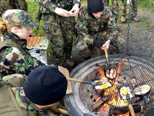 Eine Soldatin sitzt im Kreis ihrer Kameraden um ein Feuer und brät Spiegelei und Speck