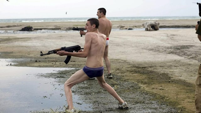 Zwei Männer in Unterhosen schiessen mit Maschinenpistolen am Strand um sich.