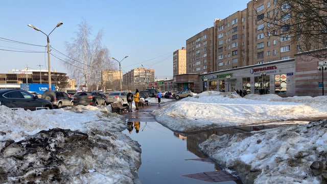 Schmutzige Schneehügel rechts und links des Gehwegs. Dahinter Menschen auf dem Trottoir, daneben braune Blockhäuser