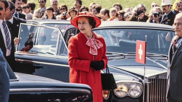 Queen in rotem Mantel und roten Hut vor einer Limousine mit Schweizerkreuz.