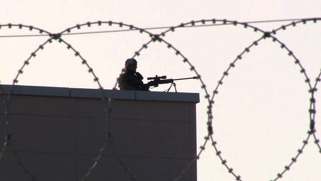 Wächter mit Scharfschützengewehr auf einem Hausdach. Stacheldraht im Vordergrund.