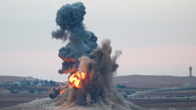 Eine Explosion mit Feuerball schleudert Erde in die Luft, im Hintergrund ist eine Stadt zu sehen.