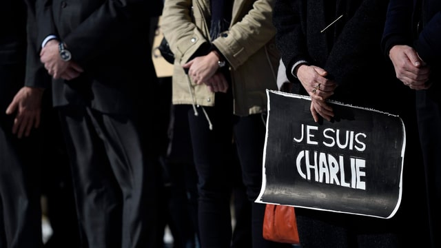 Personen mit verschränkten Händen stehen nebeneinander, eine hält ein Papier, Aufschrift «Je suis Charlie».