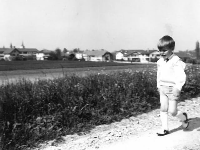 Ein kleiner Junge mit kurzen Hosen, Hemd und Pullunder läuft mit einem trotzigen Gesicht auf einem Feldweg.