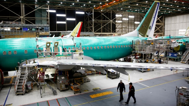 Zwei Arbeiter gehen in einem Hangar vor einem im bau befindlichen Flugzeugrumpf vorbei