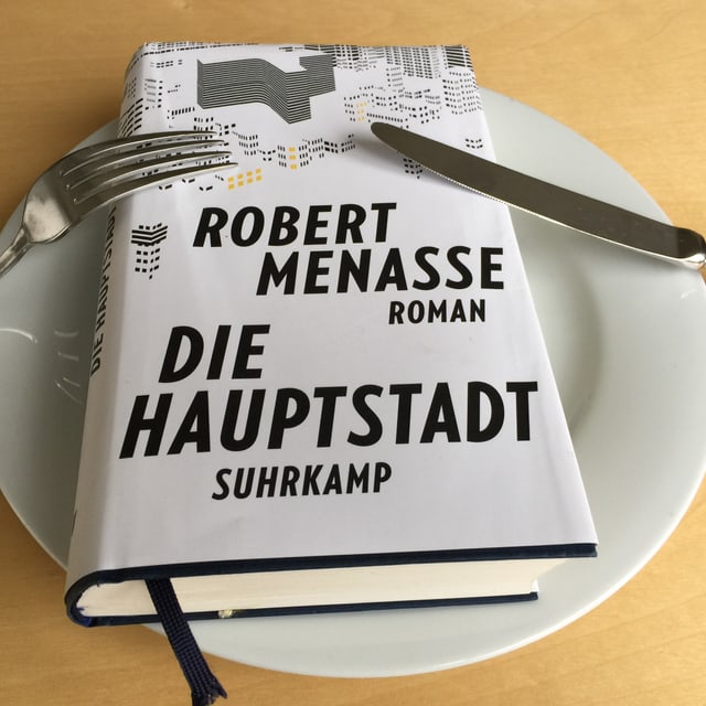 Der Roman  «Die Hauptstadt» von Robert Menasse liegt auf einem weissen Teller. Messer und Gabel angelehnt.