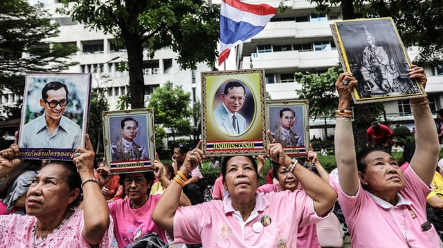 Thailänderinnen sitzen vor einem Gebäude und halten Fotos von Bhumibol in die Höhe.