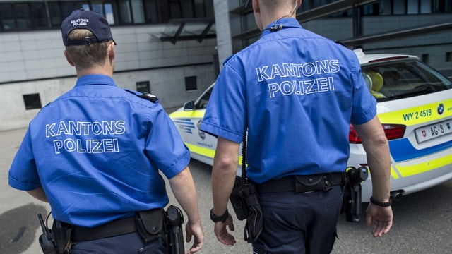 Zwei Polizisten in blauer Uniform laufen auf ein Polzeiauto zu.