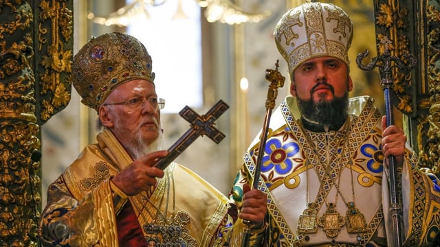 Zwei bärtige Priester in goldig verzierten Gewändern stehen in einer prunkvoll dekorierten Kirche.