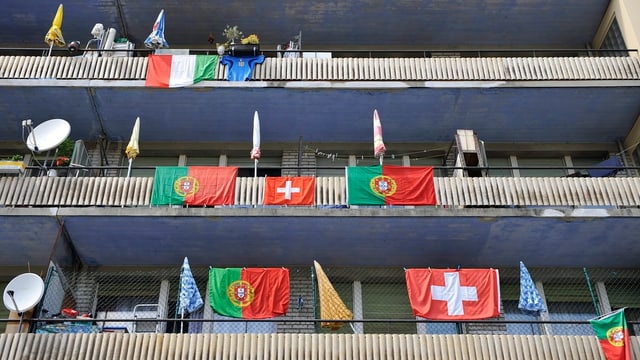 Eine mehrstöckiges Haus mit Balkonen, an denen italienische, portugiesische und Schweizer Fahnen wehen.