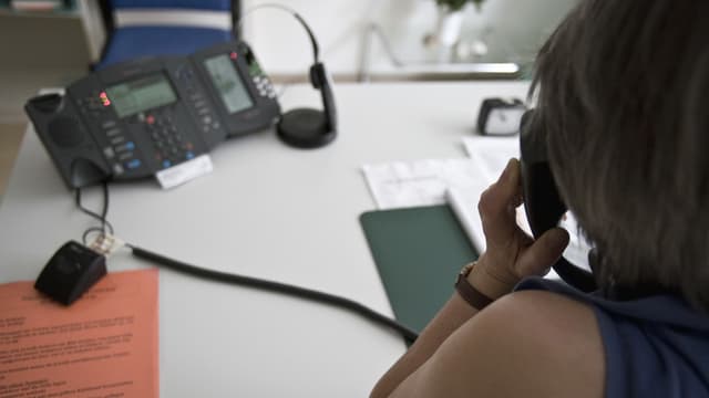 Eine Frau hält einen Telefonhörer in der Hana; sie sitzt an einem Schreibtisch.