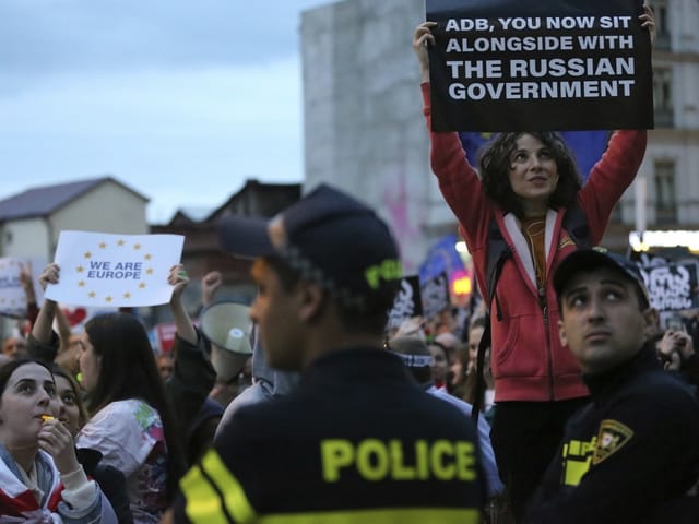 Demonstrantin hebt ein Protestplakat bei einer Kundgebung, umgeben von Polizei und anderen Demonstranten.